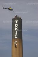 Greenpeace climbers climbing outside the chimney. They are using climbing ropes and wearing hard hats. 'Toxic' has been painted on the side. Police helicopter flying overhead.